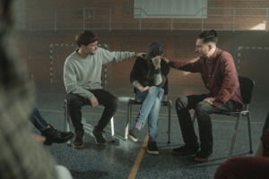 Three male youths sitting in chairs in a gymnasium. Two of the youth are consoling the youth in the middle. 