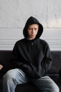 Male youth sitting down and wearing a black hoodie with the hood up. 